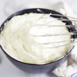 Black bowl filled with homemade whipped cream with a whisk in the bowl.