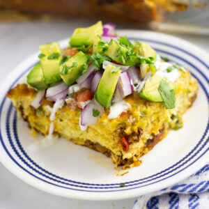 A serving of Mexican breakfast casserole on a small plate topped with fresh chopped red onions, avocado, and cilantro with a drizzle of sour cream.