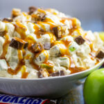 Snickers Caramel Apple Salad drizzled with caramel sauce in a large bowl next to a wrapped Snickers bar and Granny Smith Apple on a rustic wood backdrop