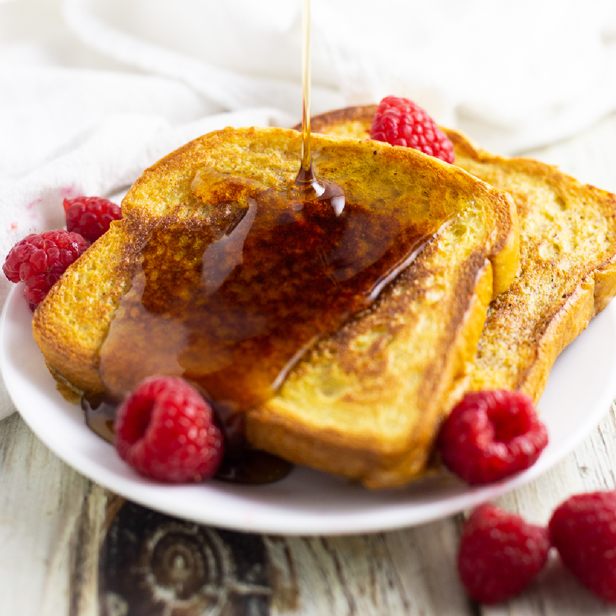 Eggnog French toast on a small white plate surrounded by fresh raspberries being drizzled in maple syrup