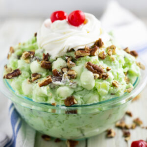 Watergate salad in a glass bowl topped with chopped pecans topped with whipped cream and two maraschino cherries.