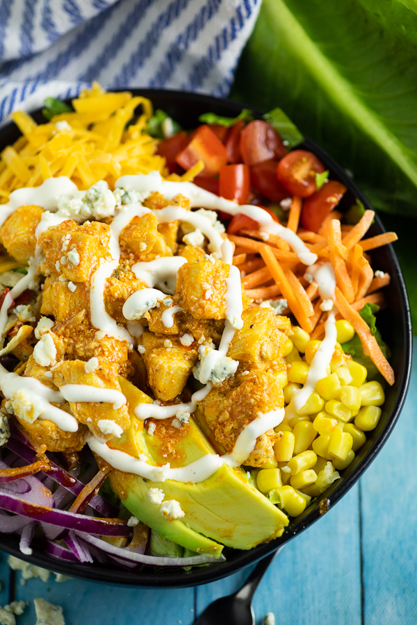 Close up of a buffalo chicken salad in a large black bowl with shredded cheese, tomatoes, shredded carrots, sweet corn, avocado slices, sliced red onions, buffalo chicken pieces, and a swirl of ranch on top