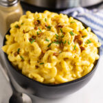 Instant pot mac and cheese in a large black bowl topped with crumbled bacon, fresh chives, and black pepper, with a black pepper shaker and Instant Pot behind it.