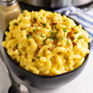 Instant pot mac and cheese in a large black bowl topped with crumbled bacon, fresh chives, and black pepper, with a black pepper shaker and Instant Pot behind it.