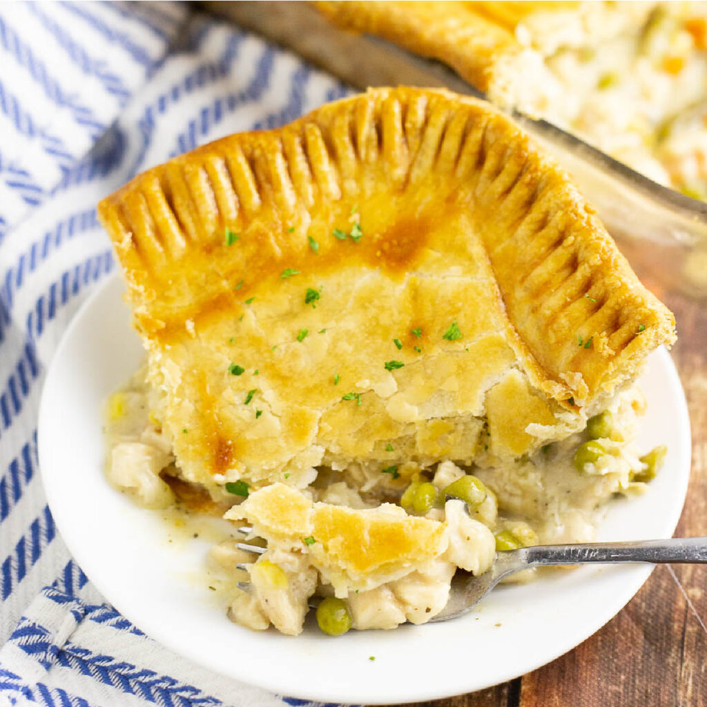 A corner slice of chicken pot pie with golden crust on a small plate with a fork with a bite on it, on a rustic wood background with a striped linen