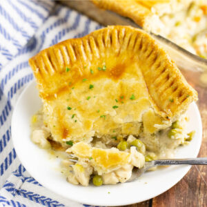 A corner slice of chicken pot pie with golden crust on a small plate with a fork with a bite on it, on a rustic wood background with a striped linen