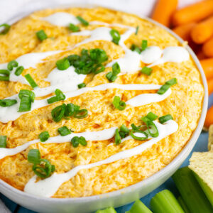 Crockpot buffalo chicken dip in a serving bowl topped with a drizzle of ranch and sliced green onions. The bowl is surrounded by carrots, celery, and tortilla chips.