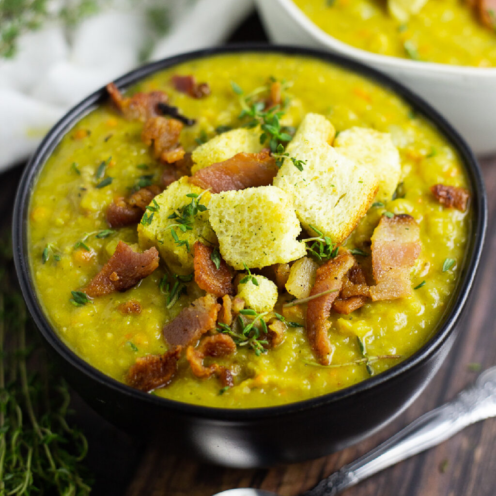 Split pea soup in a matte black bowl topped with crumbled bacon, fresh thyme, and croutons.