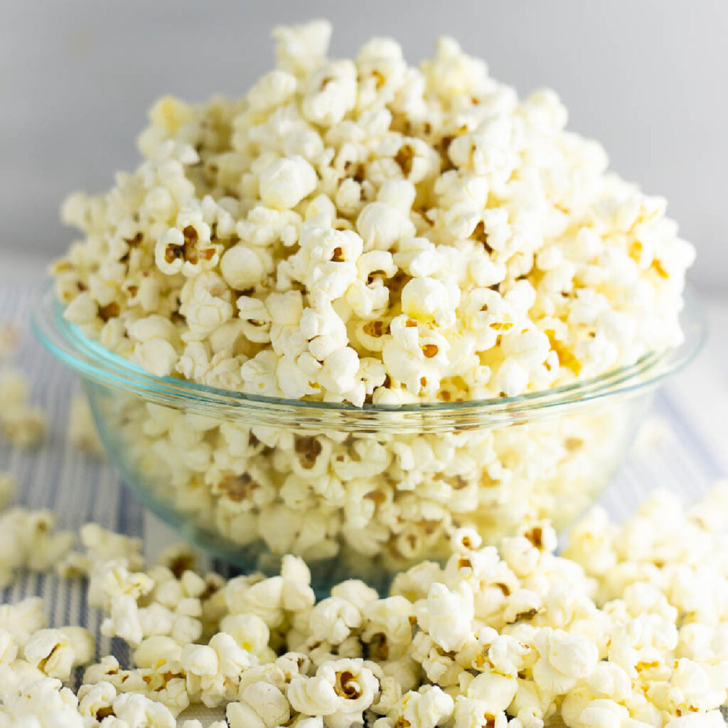 Stovetop popcorn piled high in a glass bowl with popcorn scattered all around it on a rustic white wood background.