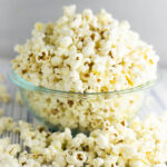 Stovetop popcorn piled high in a glass bowl with popcorn scattered all around it on a rustic white wood background.