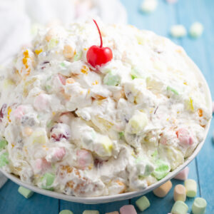 A big bowl of Ambrosia salad with a cherry on top with mini marshmallows scattered around it.
