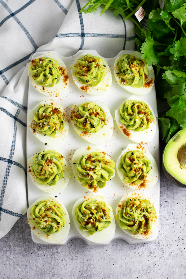 Overhead view of an egg serving tray filled with a dozen avocado deviled eggs topped with paprika and everything seasoning next to a linen napkin, half of an avocado, and a bunch of fresh cilantro