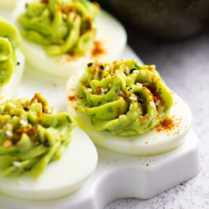 Avocado deviled eggs topped with paprika and everything seasoning on an egg serving tray next to half of an avocado and a bunch of fresh cilantro