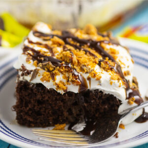 A slice of Butterfinger cake on a small blue striped place with large cake in the background with Butterfinger candy bars