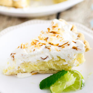 Coconut cream pie topped with toasted coconut on a small white plate next to a lime wedge and a sprig of mint.