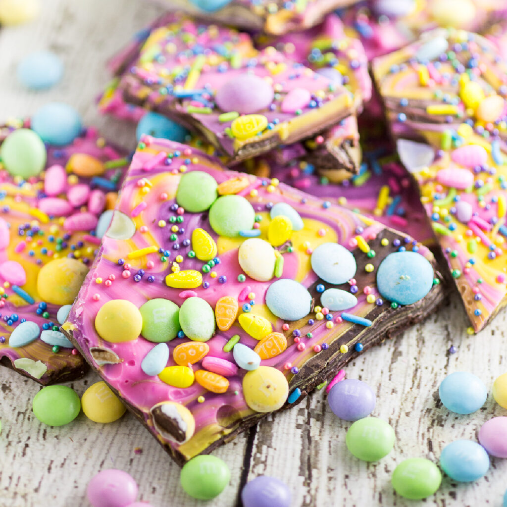 Pastel chocolate Easter bark in a pile topped with Spring sprinkles, candies, and M&M's on a rustic wood backdrop with more pastel candies scattered around.