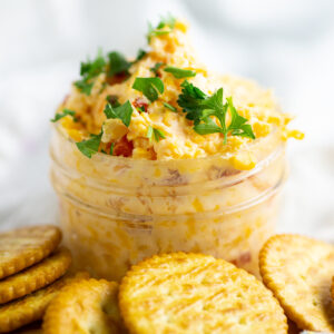 Homemade pimento cheese in a small mason jar, garnished with fresh parsley. The jar is surrounded by butter crackers.