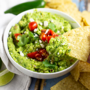 Guacamole in a bowl topped with halved cherry tomatoes, fresh cilantro, and cotija cheese with a chip dipping into and tortilla chips and a jalapeno in the background