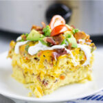 Slice of crockpot breakfast casserole on a white plate with stainless steel crock pot in the background.