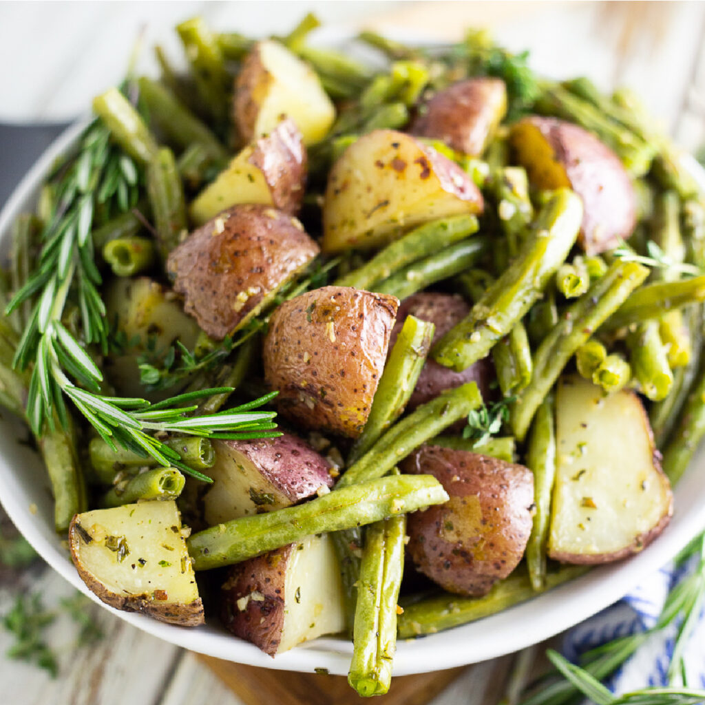 Roasted red potatoes and green beans in a white bowl with fresh rosemary sprigs on a white rustic wood background