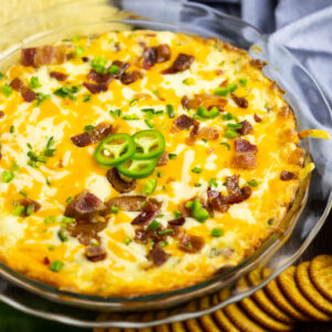 Jalapeno popper dip in a glass pie dish surrounded by chips, crackers, and a light blue linen