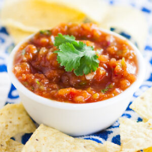 Fresh homemade salsa in a bowl topped with fresh cilantro surrounded by tortilla chips sitting on a colorful linen