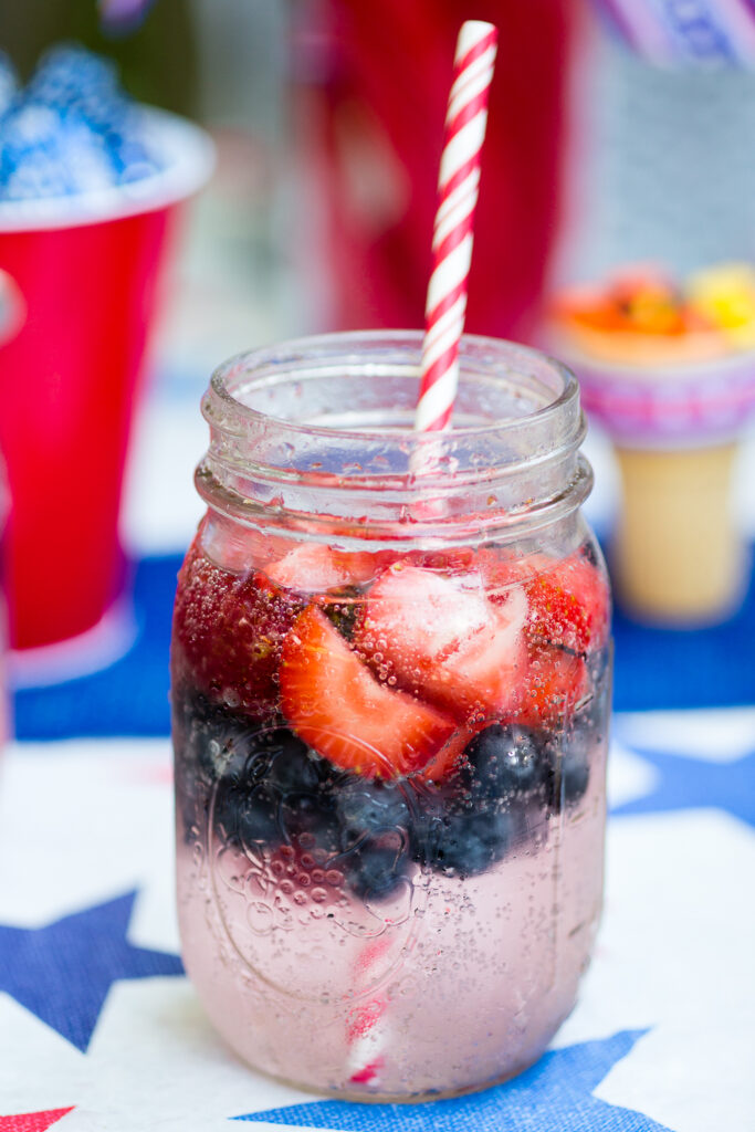 Berry spritzer in a mason jar with a layer of frozen strawberries on top and a layer of frozen blueberries under and a red striped straw in the glass