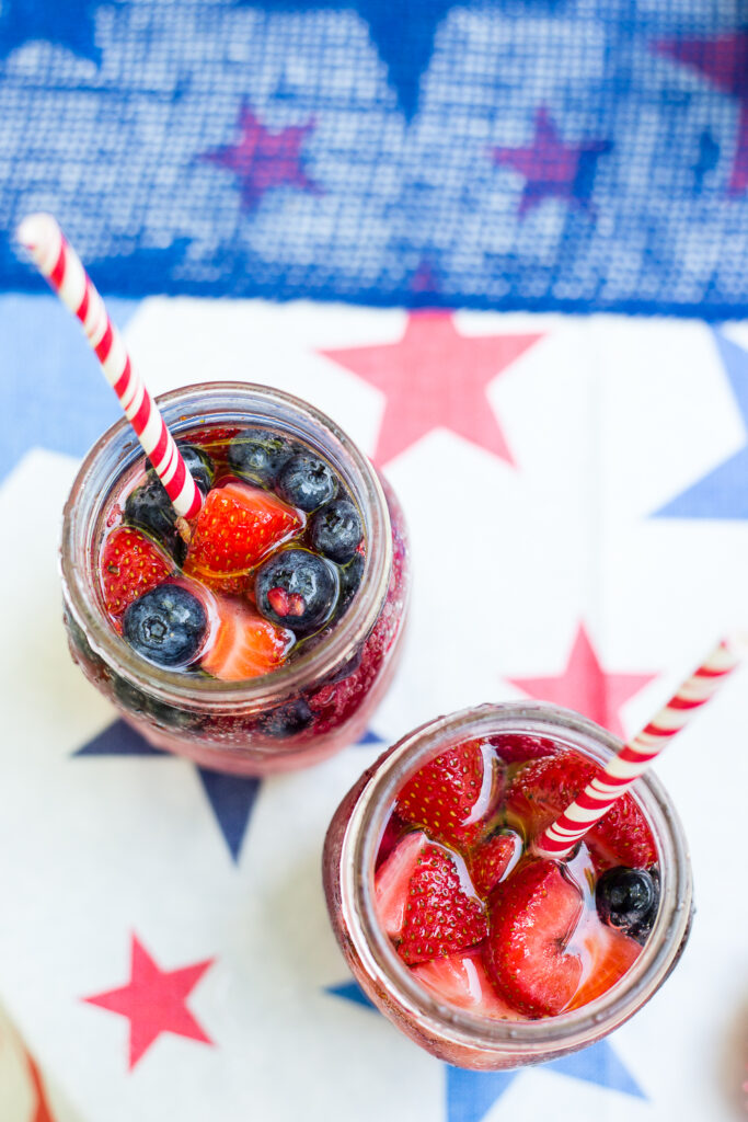 Overhead photo of 2 berry spritzers both in mason jars with red striped straws and frozen berries floating at the top.