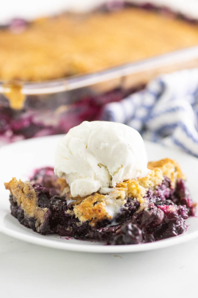 Blueberry Cobbler topped with vanilla ice cream on a small white plate and the full cobbler in the background