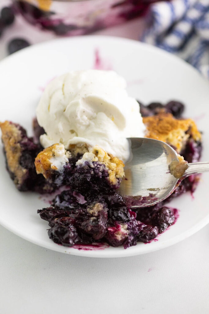 Blueberry cobbler on a small white plate with a spoon taking a bite out.