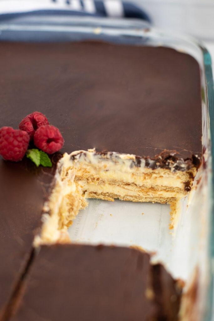 Chocolate eclair cake in a glass dish topped with raspberries with a slice missing.
