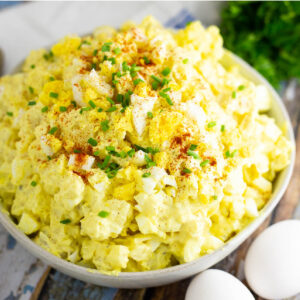 Angle shot of deviled egg potato salad in a wide bowl topped with chives and paprika, next to 2 eggs and herbs on a rustic wooden background