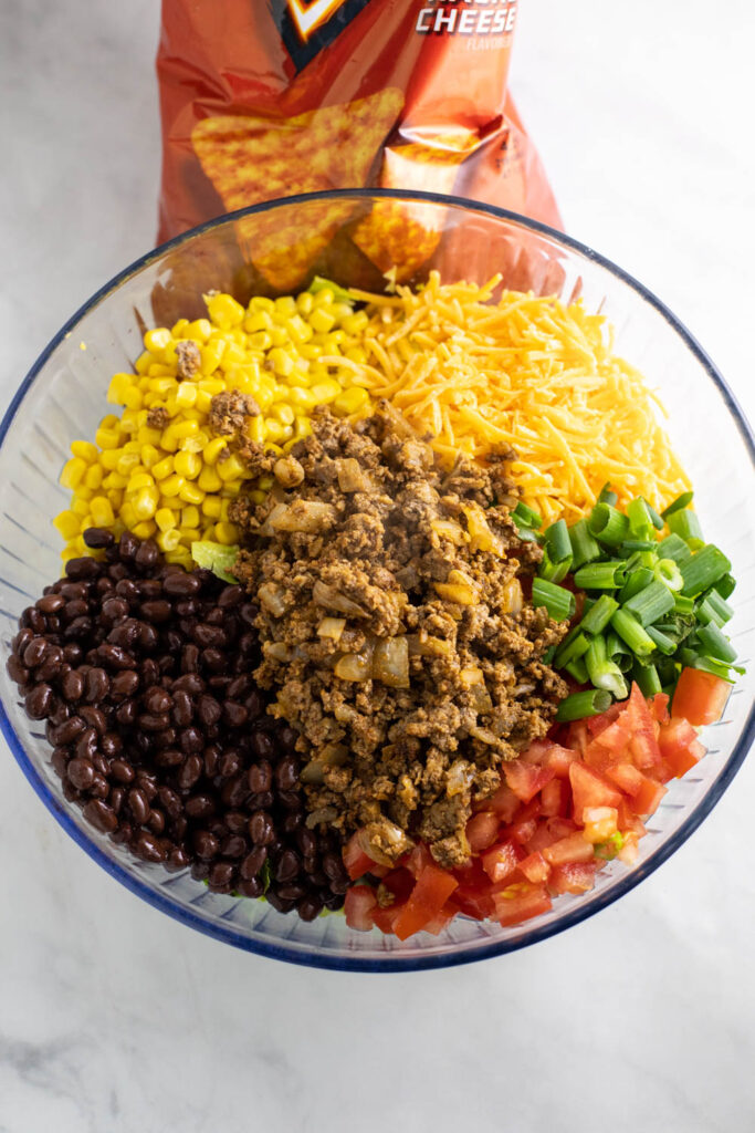 Ingredients to Dorito Taco Salad, including ground beef, black beans, corn, shredded cheese, green onions, and tomatoes, in a large glass bowl before tossing together.