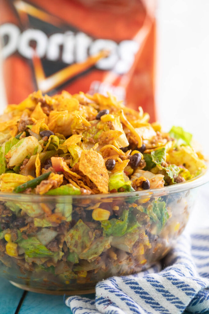 Close up side view of Doritos Taco Salad in a large glass bowl with a bag of nacho cheese Doritos behind it.