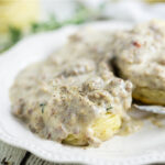 Side view of biscuits on a white plate with Southern sausage gravy dripping down, a fork on the plate, and more biscuits and thyme behind them