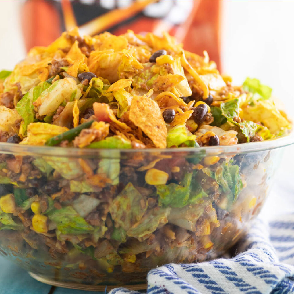 Close up side view of Doritos Taco Salad in a large glass bowl with a bag of nacho cheese Doritos behind it.