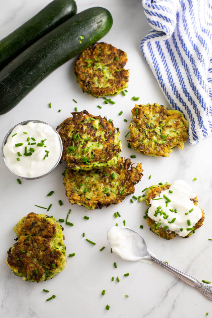 Zucchini fritters scattered on a white marble countertop with sour cream and fresh chopped chives and two whole zucchini