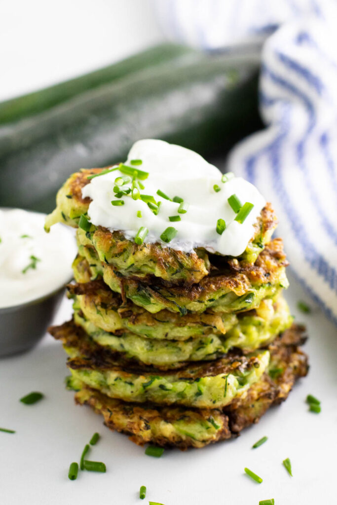 Zucchini fritters in a stack topped with sour cream and fresh chives with a small cup of sour cream and a zucchini in the background.