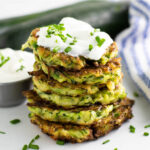 Zucchini fritters in a stack topped with sour cream and fresh chives with a small cup of sour cream and a zucchini in the background.