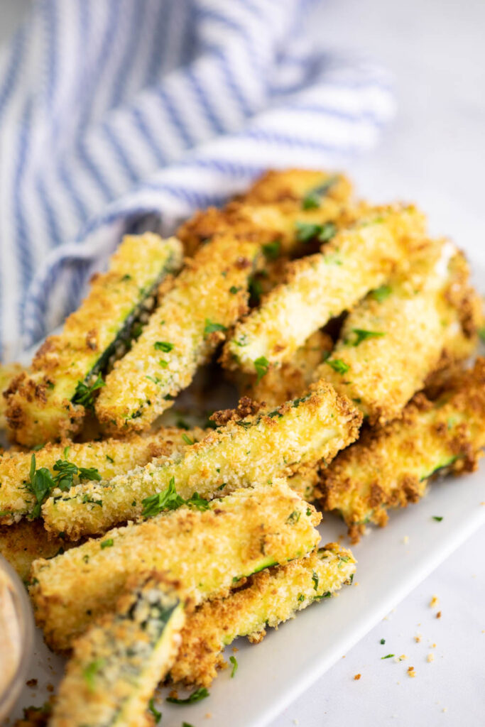 Pile of air fryer zucchini fries on a white rectangle plate in front of a striped linen on a marble backdrop