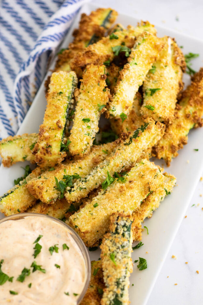 Rectangle plate filled with air fryer zucchini fries next to sauce topped with fresh chopped parsley