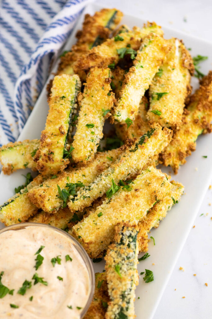 Rectangle plate filled with air fryer zucchini fries next to sauce topped with fresh chopped parsley