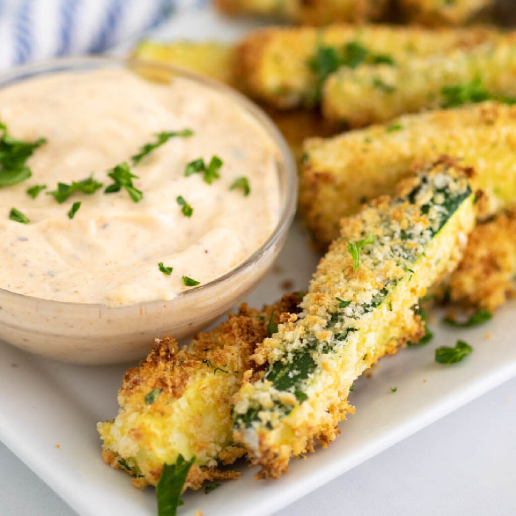 Air fryer zucchini fries sitting next to a sauce in a glass bowl topped with chopped fresh parsley