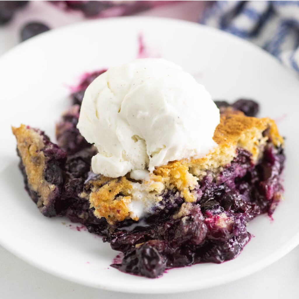 Blueberry Cobbler topped with vanilla ice cream on a small white plate and the full cobbler in the background