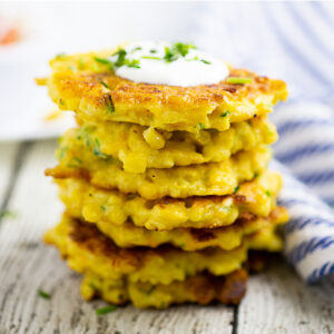 Stack of corn fritters topped with sour cream and freshly chopped chives on a rustic wood background.