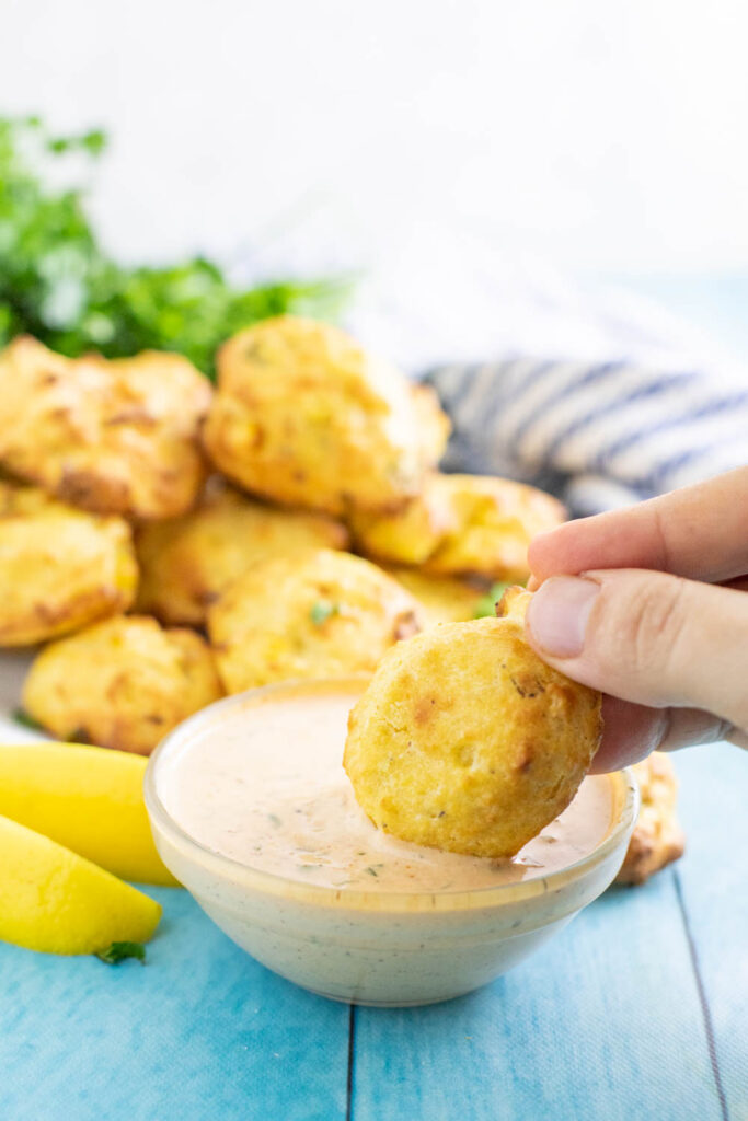 Air Fryer Hush Puppy dipping into a small glass bowl of remoulade. The bowl is sitting in front of 2 lemon wedges and a pile of air fryer hush puppies