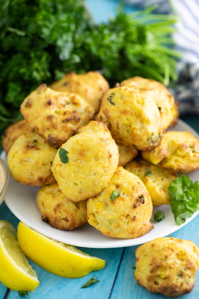 Small plate piled with air fryer hush puppies next to 2 lemon wedges with a bunch of parsley in the background