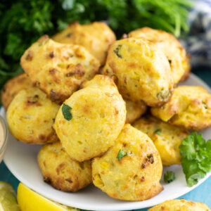 Small plate piled with air fryer hush puppies next to 2 lemon wedges with a bunch of parsley in the background