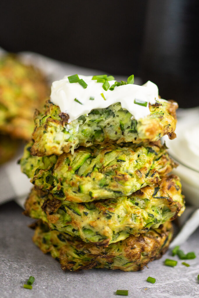 Stack of 4 air fryer zucchini fritters on a concrete background with a white linen. The top fritter is topped with sour cream and chives and there's a bite taken out
