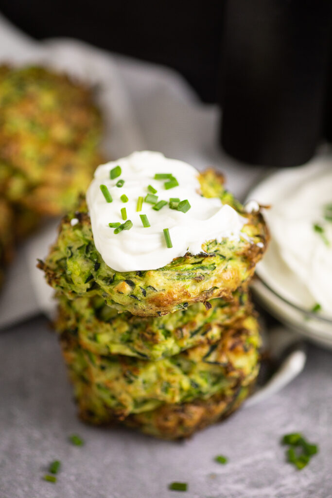 Stack of 4 zucchini fritters on a concrete backdrop with chives scattered all around. The top fritter is topped with sour cream and chopped chives.